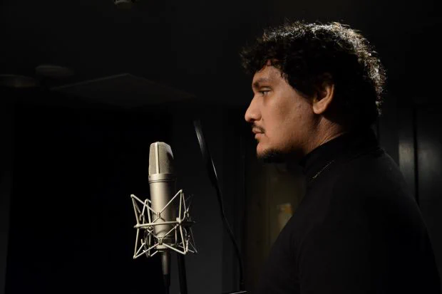 A man with black hair and moustache stands in the dark at a microphone. He is wearing black.