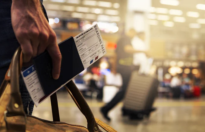 Primer plano tenemos la mano de una persona sujetando unos boletos de avion y pasaporte, además está cargando una maleta de mano y en segundo plano personas en un aeropuerto. 