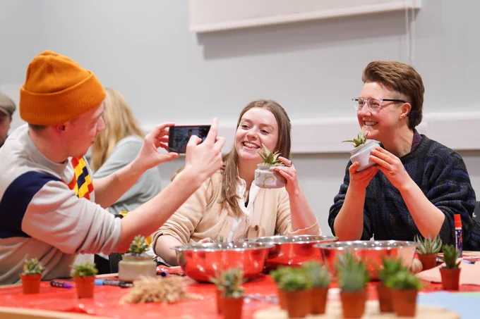 a man takes a photo using his phone of two smiling people holding cactuses