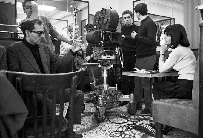 A monochrome photograph captures the director Jean-Luc Godard conversing with actress Chantal Goya, who is seated at a table on a film set. Meanwhile, the film crew talks behind the camera, which is directed at Goya.