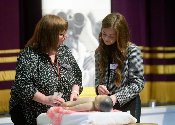 two women stand on stage demonstrating baby massage to an audience.