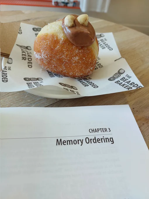 Chocolate filled doughnut with hazelnuts on top, resting on a plate next to the corner of a book, with the page showing 'Chapter 3 - Memory Ordering'