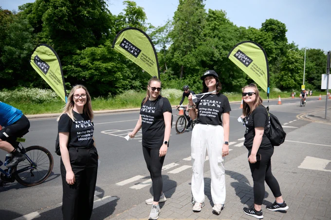 members of the Anthony Nolan team cheering our cyclists on, wearing Anthony Nolan branded t-shirts, and holding large green Anthony Nolan flags