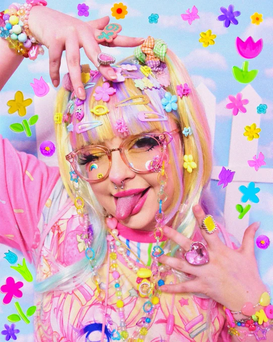 A studio photograph of Pixie (Jill & Sunny fronting) in a full decora look. They're wearing a pastel rainbow wig with blunt bangs and two half-up twintails absolutely packed with pastel hairclips, stickers on their face, glasses with a pastel glitter rainbow chain, several stacked pastel rainbow necklaces and an over-sized Ocean in Space tee. Rings and bracelets are also stacked on in excess. They pose in front of a blue sky wallpaper backdrop with a picket fence prop and have edited gel window cling flower images into the edges of the photo.