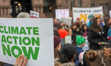 An image of a gathering of folks, in the bottom left is a sign "Climate Action Now" and other signs indicating action on the climate crisis can be made out in the background