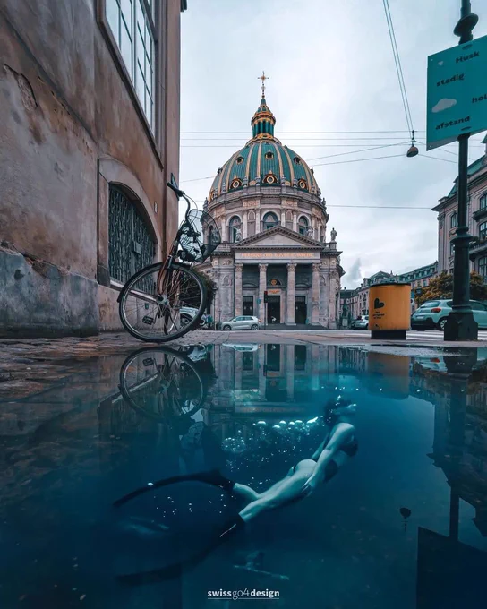 A street in an old town with a structure in the background. There's a large puddle on the sidewalk with a diver swimming to the surface.