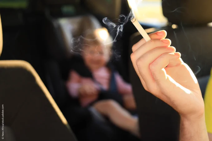 Closeup of a hand holding a cigarette in car and in a while a child is sitting on the back seat. 