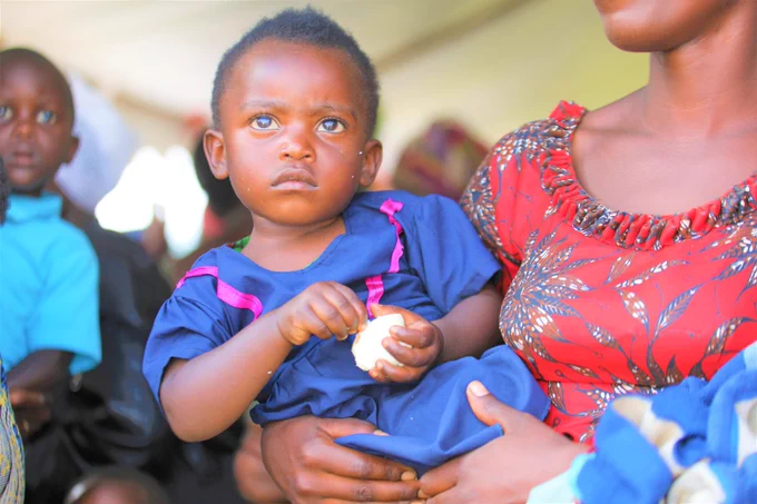 A child, carried by her mother, holds a peeled egg in her hands.