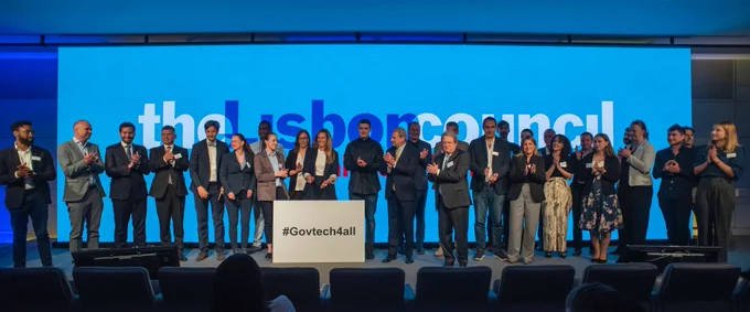40 people clapping on a stage with a panel reading “#GovTech4All”