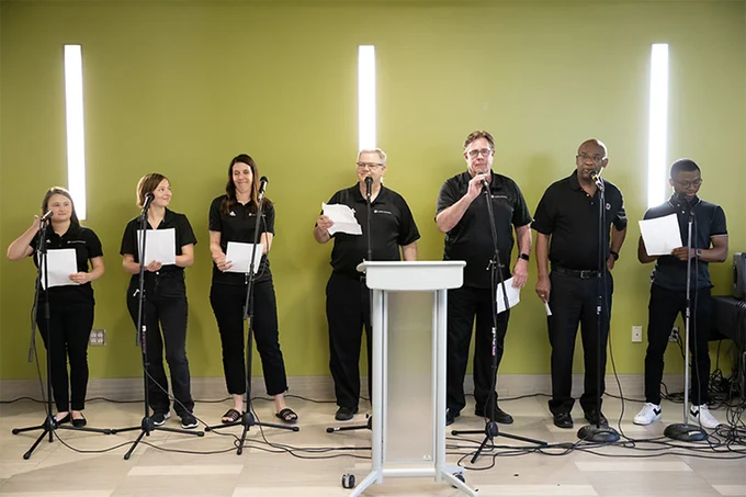 Seven people stand in a sunny atrium in front of microphones, ready to perform a song