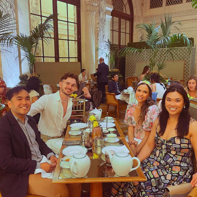 There is a man wearing a pearl necklace along with a white shirt, a man wearing a dark grey blazer, a woman wearing a floral dress and headscarf, and a woman wearing a dress depicting an assortment of sardine can artwork. All four people sit at a dining table. The table has a spread of dishes, tea cups, glasses, utensils, and teapots. Multiple tables, chairs, and restaurant guests are present in the background. There are also two sets of double French doors and three potted palm plants with floral arrangements in the background.