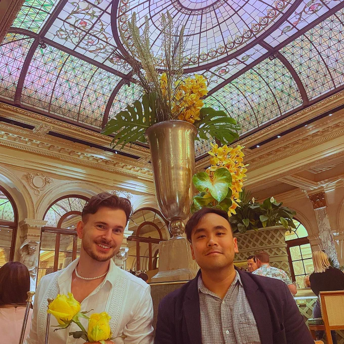 A man wearing a pearl necklace along with a white shirt and a man wearing a dark grey blazer sit a dining table. Behind them is a potted floral arrangement with palm leaves. There is also a stained glass ceiling in the background.