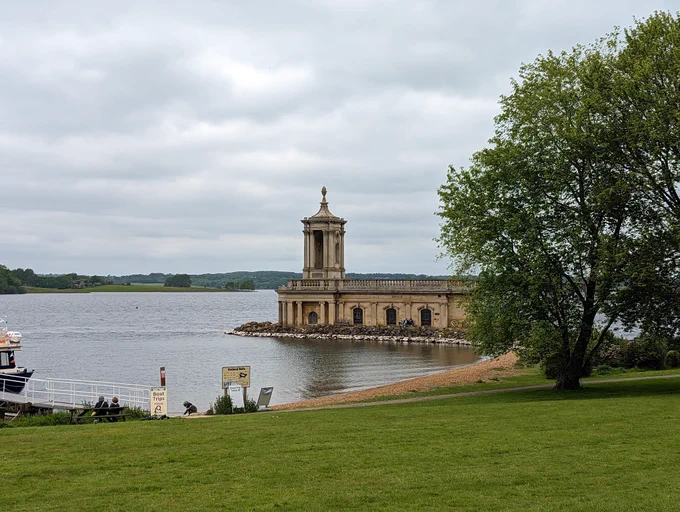 Photo of a small yellow stone church with a little tower standing on a jetty on a lake. There is a grassy field with a tree in front, and fields and woods visible on the other side of the lake. A boat is just visible on the left of the photo.