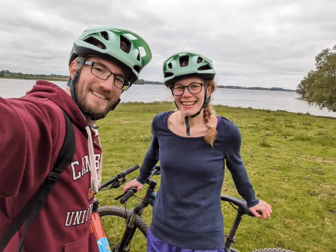 Photo of two people smiling wearing cycling helmets standing on some grass in front of a big lake. The sky is grey and cloudy and the people are wearing sweaters.