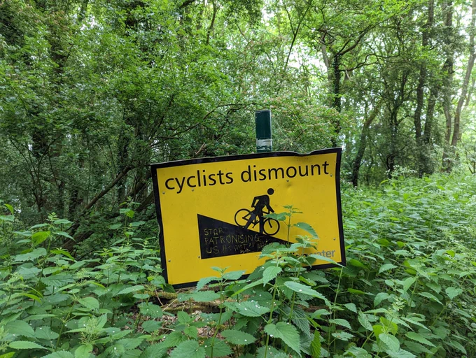 Photo of a big yellow sign among some nettles and trees. The sign has a picture of a slope with a person walking next to a bike and the text ‘cyclists dismount’. Onto the slope someone has scratched the words ‘stop patronising us!!’ And then some more words that are more difficult to make out but might be meant to say ‘we know what we all want’ (to cycle down the hills)