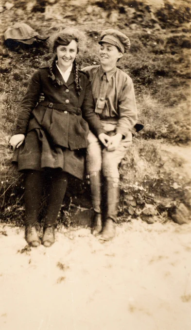 Two young women sitting on a grassy bank, holding hands and laughing. Alice is wearing a collared shirt, pants, and chauffeur’s cap.
