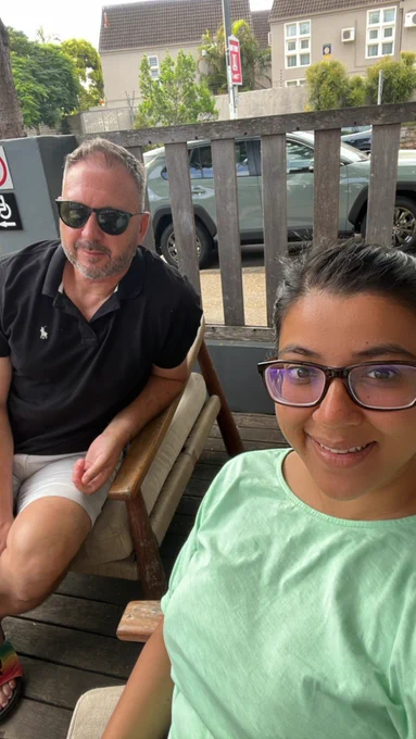 Robert Hillier, a white male in his early 50s wearing sunglasses, shorts and a black polo shirt sits behind Feyaza Khan, who is (and looks) ethnically Indian wearing a light green T shirt and square glasses. They’re both looking at the camera and a wooden fence is visible behind them with a car parked behind it. Also visible in the image is brown decking and comfortable wood and beige chairs.