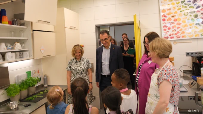 Bundesminister Cem Özdemir und Ministerin Katrin Eder sprechen mit Kitakindern. Sie stehen in einer Kinderküche.