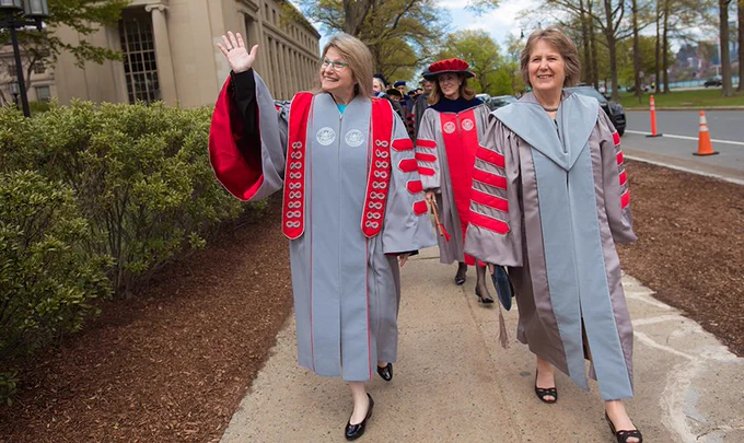 Sally Kornbluth walks next to Diane Greene and in front of Cindy Barnhart and other MIT faculty along Memorial Drive, all wearing academic regalia. Kornbluth, Greene, and Barnhart all wear cardinal-and-silver MIT robes.