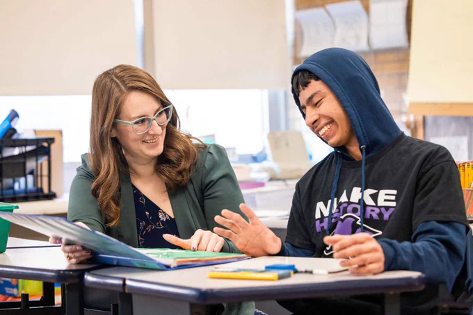 A woman and a boy sit next to each other at desks in a classroom. They smile as she points to a sheet of paper in a three-ring binder that she has opened.