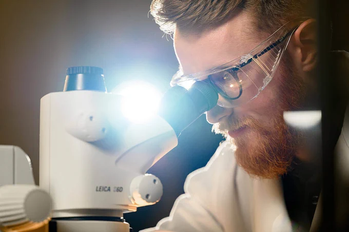 A scientist wearing a white lab coat and safety goggles peers through a microscope.