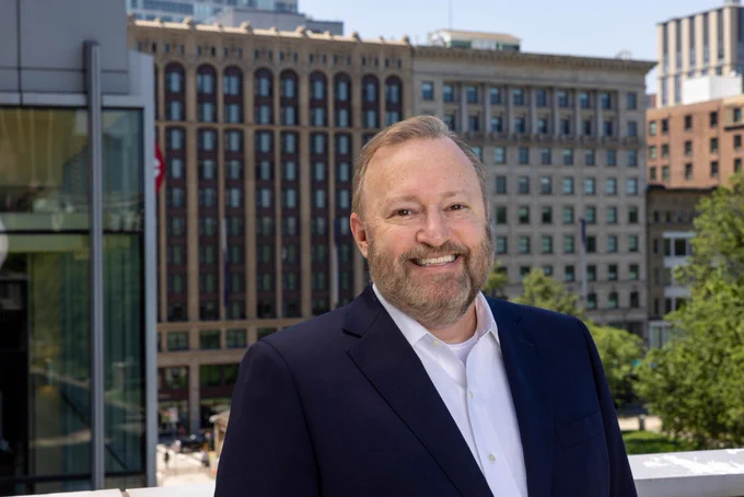 Smiling man in suit jacket and shirt with large building in the background behind him.