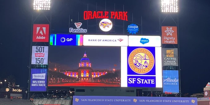The scoreboard screen at Oracle Park displays a photo of San Francisco City Hall at night with purple and gold lights
