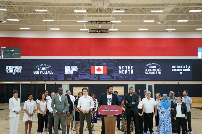 A man is standing at a podium and speaking. Prime Minister Justin Trudeau, Cabinet ministers, Members of Parliament, Masai Ujiri, and other people are standing behind him.