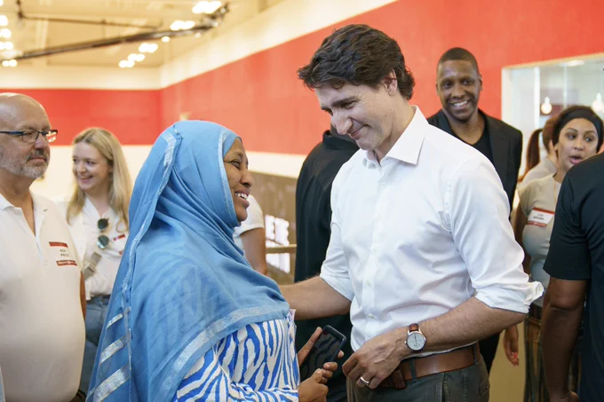 Prime Minister Justin Trudeau is standing and speaking with a woman. Both of them are smiling. Other people, including Masai Ujiri, are standing behind them.