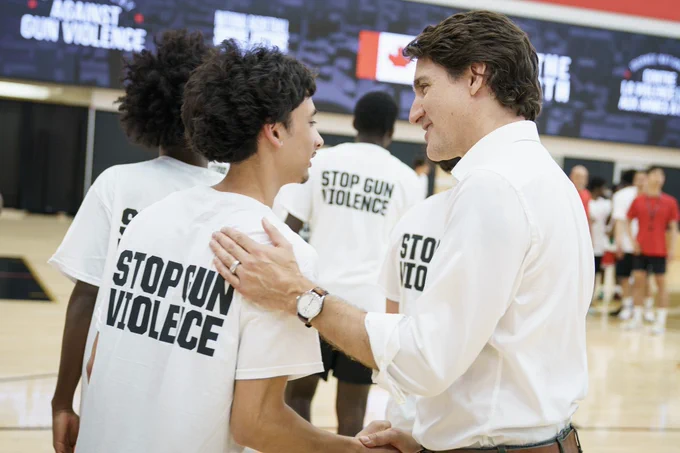 Prime Minister Justin Trudeau is standing and shaking hands with a young man. The Prime Minister’s hand is on the man’s shoulder. Both of them are smiling. Other people are standing behind them.