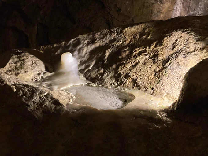 A dimly-lit photograph of Lake Inferior inside Crystal Cave. Lake Inferior is a puddle of water a few inches deep and located in the middle of the photo. It is surrounded by rocks on all sides, and the photo transitions into darkness on all sides. 