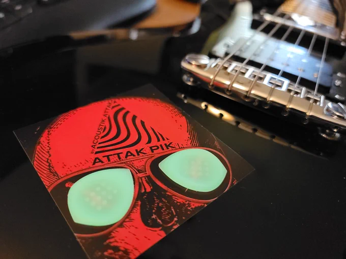 A sticker with top half of a red skull graphic featuring logo of Attak Pik placed on a black PRS guitar. There are also 2 Radium glow-in-the-dark picks placed over the mirrored shades the skull is wearing. 