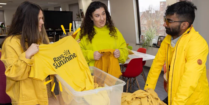 A group of three people in an office. They are stood around a box of yellow clothes, and are also wearing all yellow. 