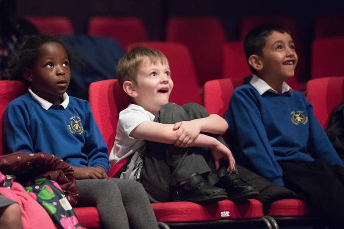 Three children in school uniform sit in a theatre with expressions of awe on their faces