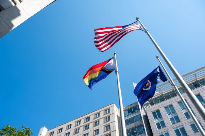 A view from the ground up at the rainbow  Progress Pride flag is seen flying at the Mary W. Jackson NASA Headquarters Building, Thursday, June 1, 2023, in Washington. In the image, it's between the U.S. flag and a flag carrying the NASA insignia. The background is a cloudless blue sky. Credit: NASA/Keegan Barber