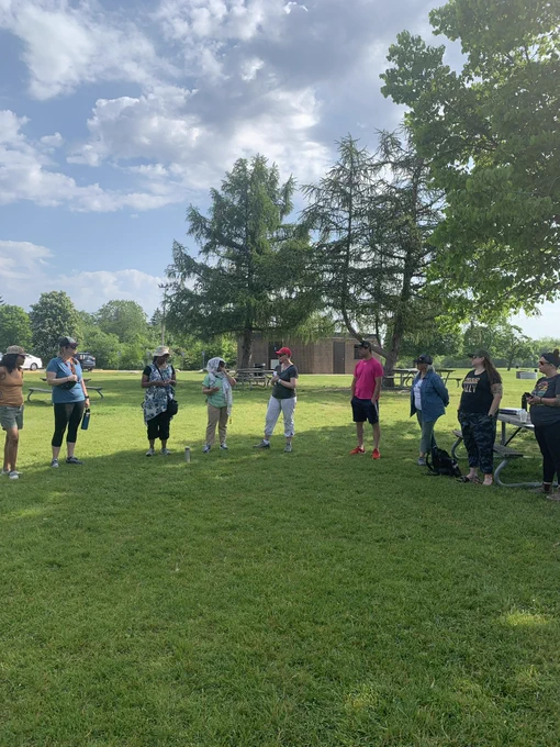 Nine adults standing in a semi-circle outdoors. Surrounded by trees and open green space.