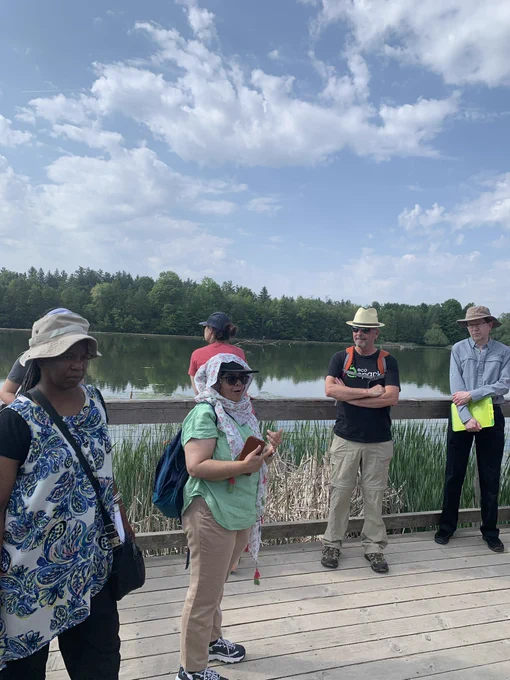 Six adults outdoors standing on a wooden boardwalk  overlooking water that is surrounded by trees and marsh. Two of the adults are facing in the direction of the water while the other four are facing the camera.