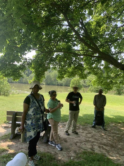 Four adults standing outdoors below a tree and in front of a bench. A lake is behind them in the distance.