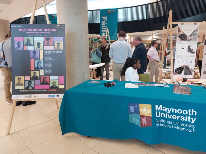 Image of people at a showcase event. There is a table covered by a table cloth with Maynooth University logo on it. There is a large poster with information about the event. 
