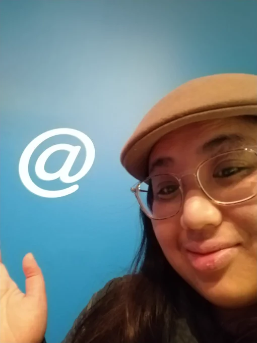 Kawa is wearing a brown flat cap and clear glasses. They are gesturing next to the Ray Tomlinson @ sign on display at the "Never Alone" NY Museum of Modern Art exhibit, much like their Twitter icon.