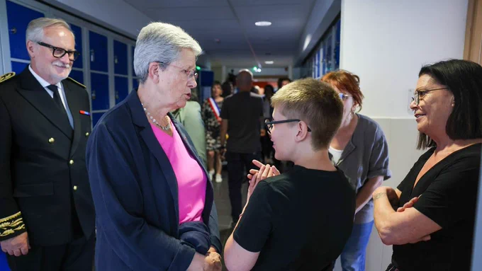 Geneviève Darrieussecq discute avec un jeune élève dans les couloirs de l'école. Autour d'eux, des adultes écoutent leur discussion. En arrière-plan, des casiers de couleurs bleue.