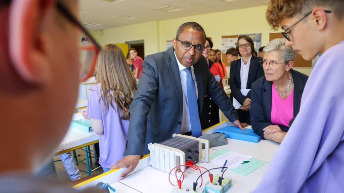 Pap Ndiaye et Geneviève Darrieussecq sont dans une classe avec deux  élèves et de l'animation autour d'eux. Ils sont appuyés sur une table dédiée aux travaux pratiques. Les ministres échangent avec deux enfants au premier plan.