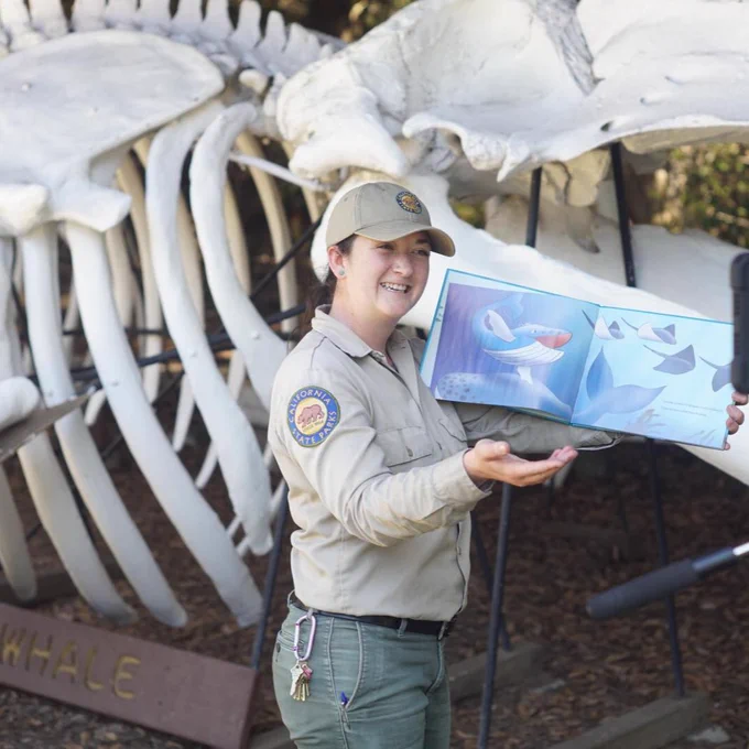 State Parks Employee holding a children's book open. We see drawings of a whale with multiple sting rays. She is standing in from of a large whale skeleton.
