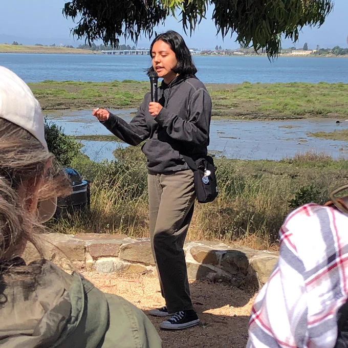 Irma Mendoza Gomez, a young Latina adult wearing a grey sweatshirt and brown pants, gestures as she speaks into a microphone to a group of people along the Oakland shoreline.