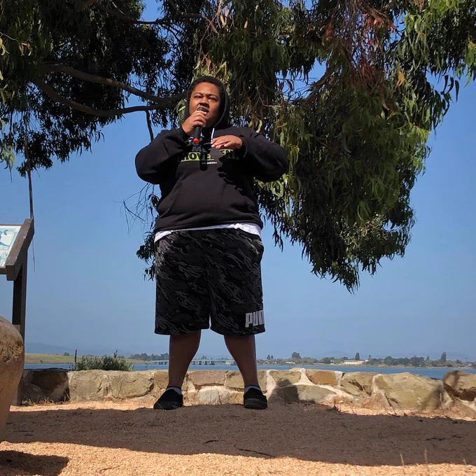 Shy Walker, a young Black adult in a black hoodie and shorts, speaks into a microphone while standing under drooping tree branches along the Oakland shoreline.