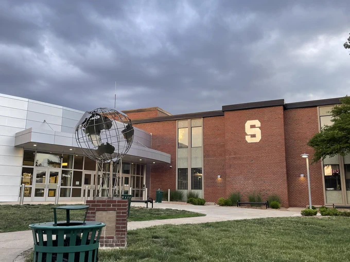 The main entrance of Shawnee Mission South High School at dusk. In the foreground, the globe sculpture. On the side of the building, the large metallic S reflects the setting sun. Behind the building, brooding clouds begin to gather.