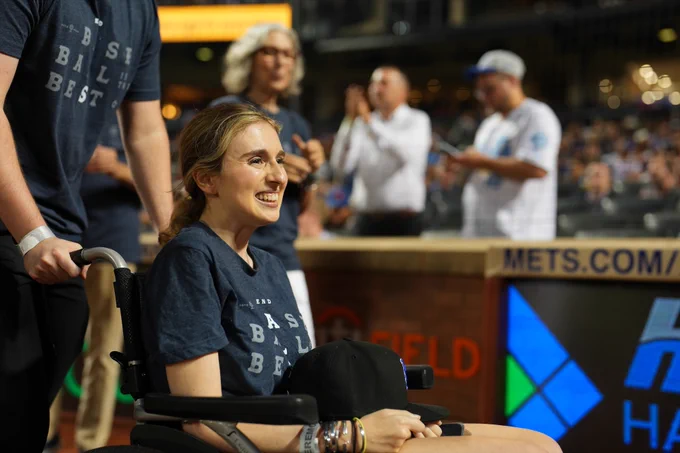 Sarah Langs smiles as she is honored at Citi Field Friday night on Lou Gehrig Day. She wears a gray "Baseball is the Best/End ALS" t-shirt and holds a black Mets cap in her lap.
