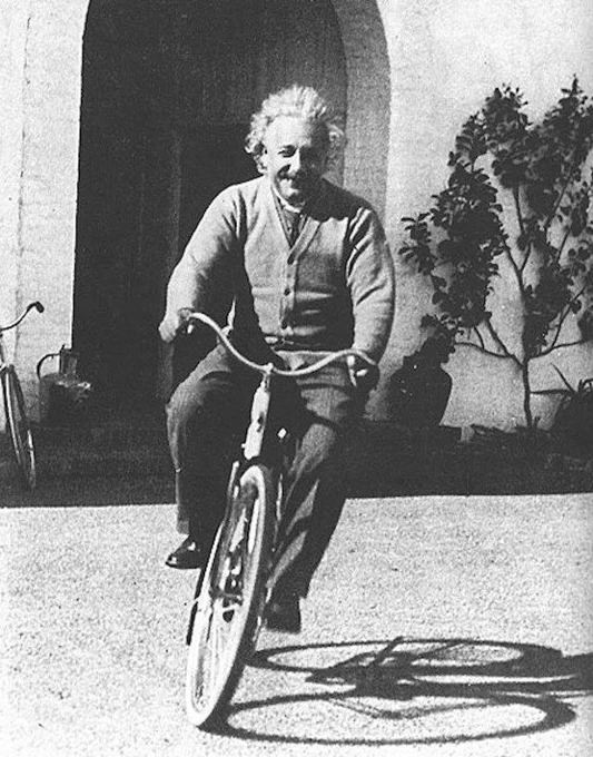 Black and white photo of a happy Albert Einstein riding a bicycle.