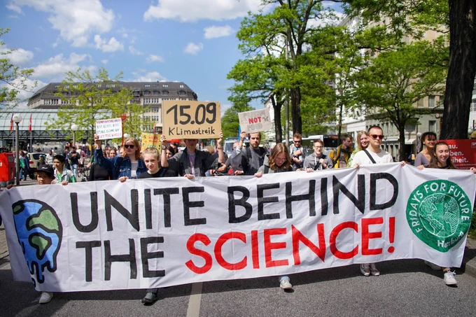 Auf dem Frontbanner einer Klimademo steht Unite behind the Science. Eine Person hält ein Demoschild mit der Aufschrift 15.09. Klimastreik