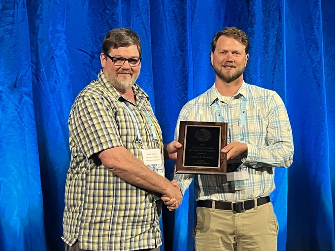 Two men in front of a blue background, one is receiving an award